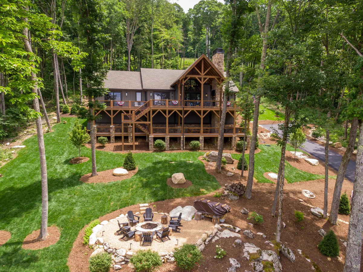 The image showcases the inviting wooden exterior of a luxurious single-level cabin nestled in a lush, green mountain landscape. The centerpiece is a beautifully arranged fire pit area surrounded by Adirondack chairs, perfect for gatherings. The home is enveloped by mature trees, creating a sense of privacy and peacefulness, while hints of mountain views peek through the foliage.