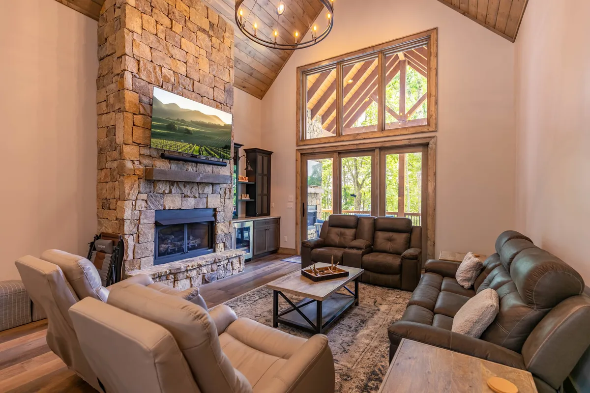 A comfortable great room scene featuring a striking stone fireplace, plush leather seating, a large flatscreen TV, and expansive windows framing serene views. The ambiance is cozy and perfect for relaxing or hosting special gatherings.
