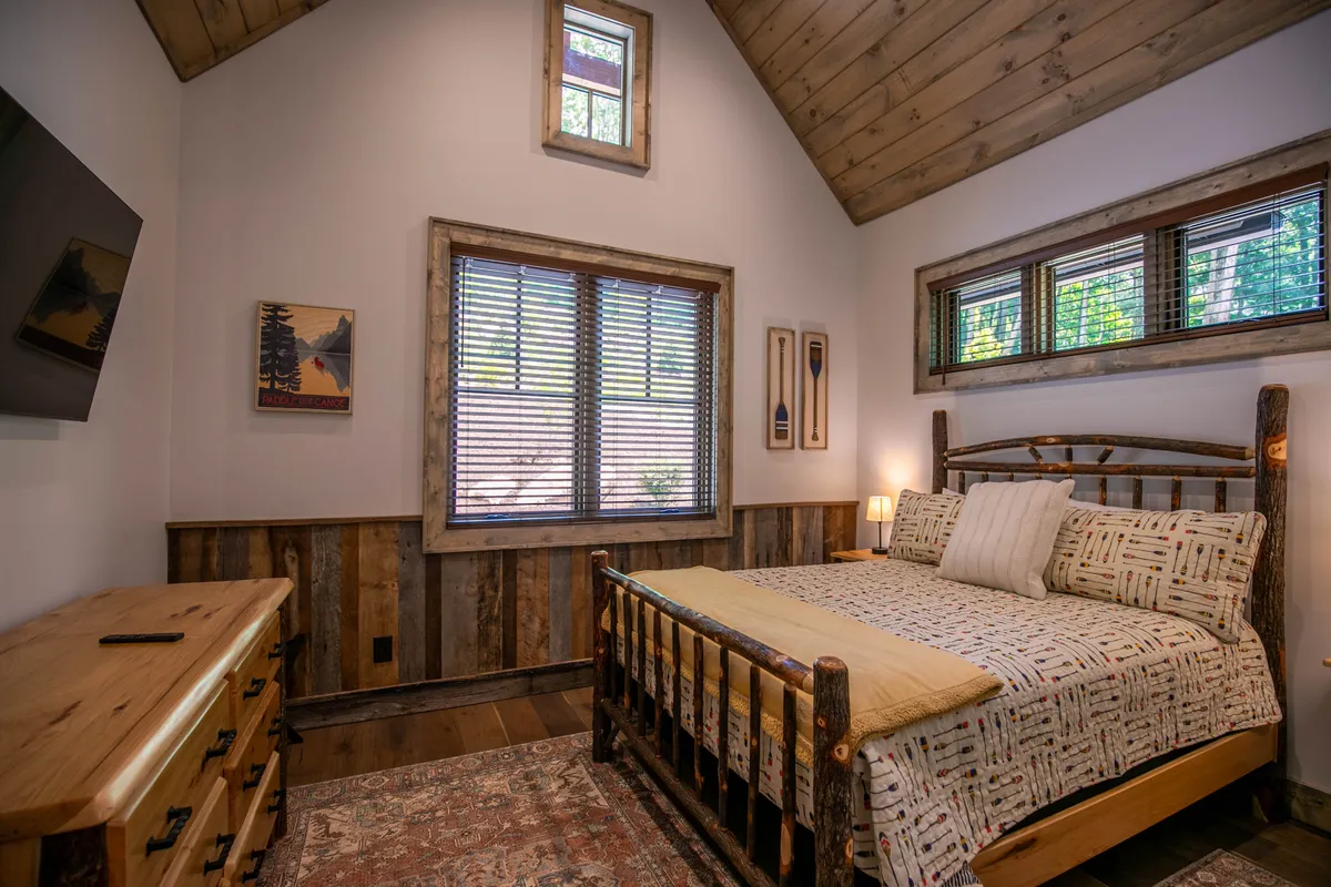 A tranquil bedroom featuring a cozy log bed, window with blinds allowing natural light to flow in, wooden dresser, and carefully chosen wall art. The calming decor and plush bedding create a restful retreat, ideal for unwinding after a day of adventure.