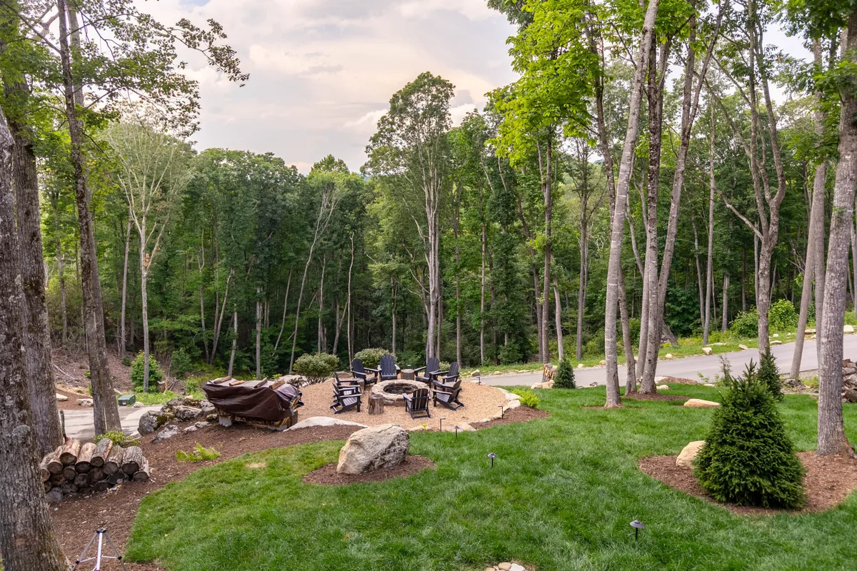 A close-up of the fire pit area, with surrounding chairs and a wood pile nestled in natural landscaping. The tranquil setting is ideal for intimate gatherings, sunset views through the trees, and early morning coffee in nature.