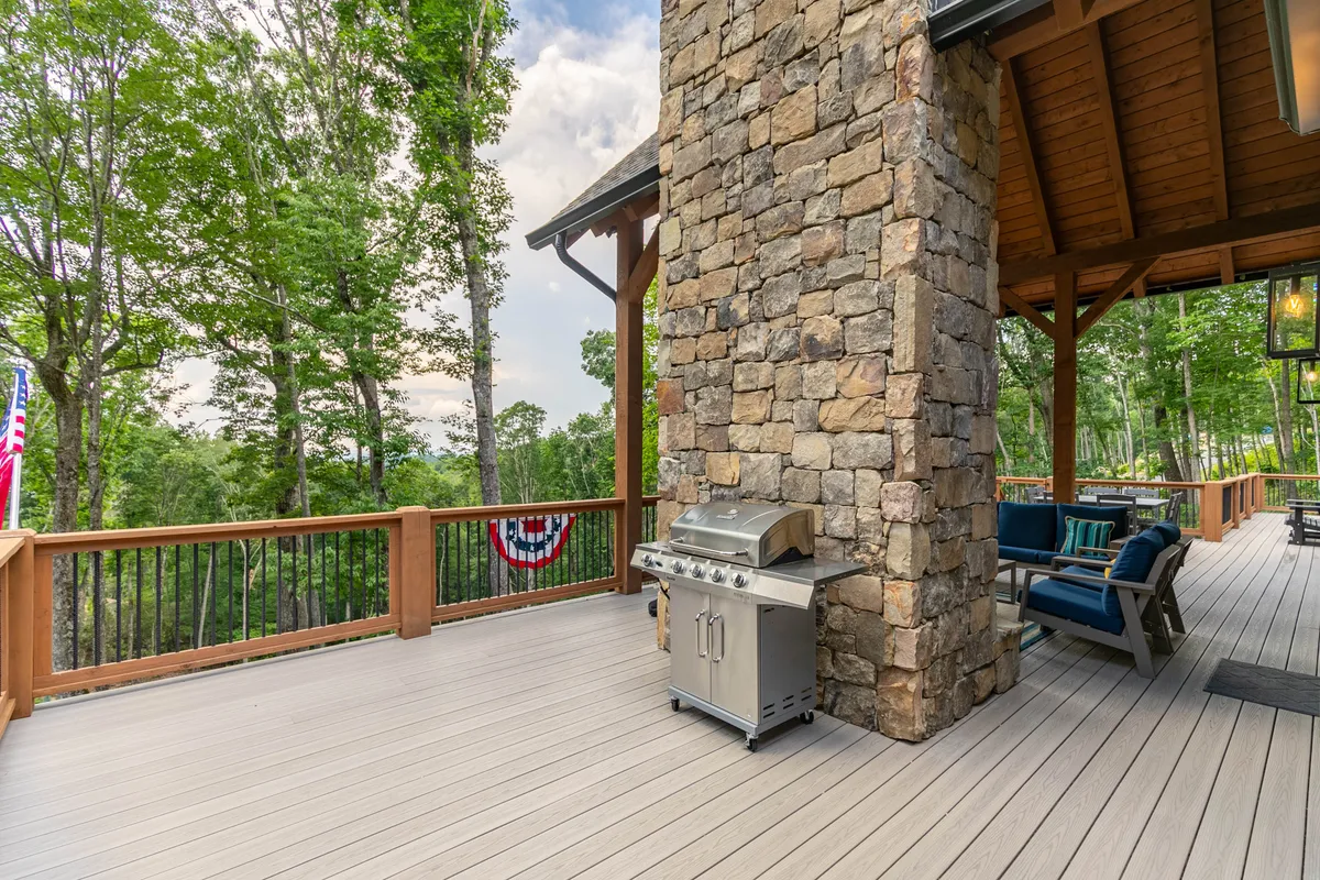 An exterior shot showcasing a well-equipped grill and adjacent stone wall on the deck, complemented by a comfortable seating arrangement and a backdrop of mature trees. The scene suggests a space perfect for barbecues, social gatherings, and unwinding outdoors with scenic views.