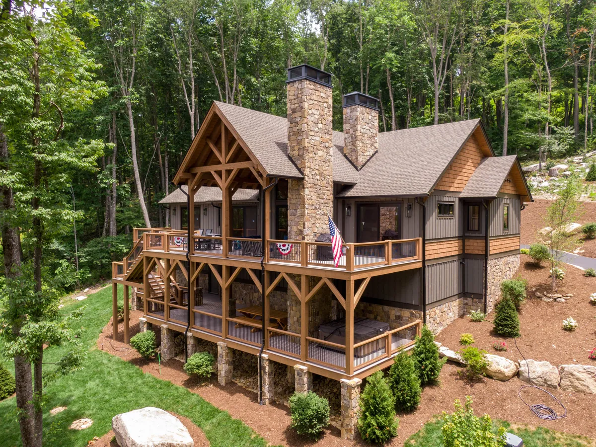 The exterior view showcases a warm, inviting wooden home with a large deck, stone chimney, and front porch. The large windows hint at bright, open indoor spaces, while the natural setting encourages outdoor gatherings and exploration.