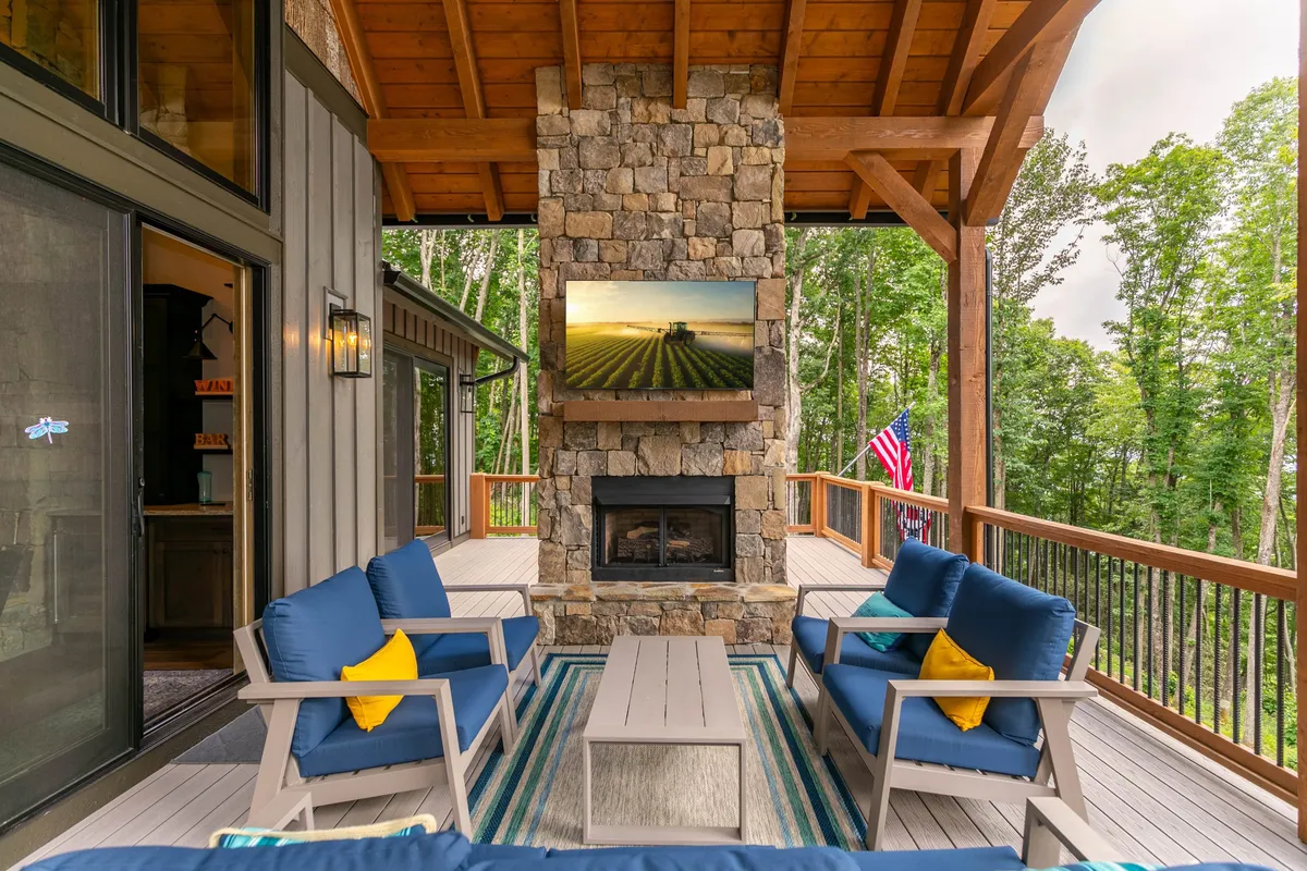 This image showcases a covered deck furnished with a stone fireplace, comfortable outdoor seating, and a mounted TV. Decorative pillows and a scenic view complement the inviting atmosphere, ideal for al fresco dining or relaxing by the fire.