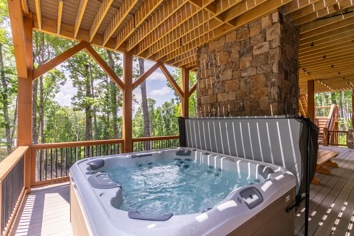 A serene wooden deck with a private hot tub is nestled among trees, bordered by a stone wall. The setting evokes tranquility and offers a rejuvenating spot for soaking while enjoying surrounding nature and mountain views.
