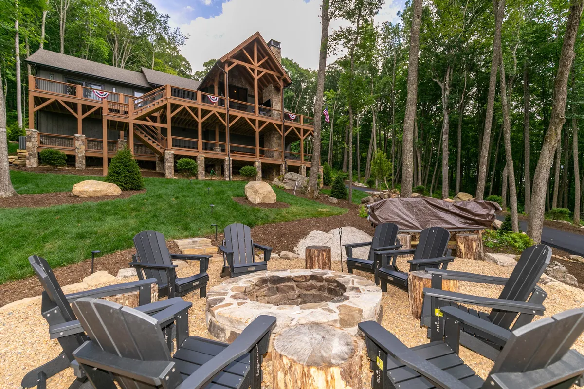A spacious wooden deck provides ample space for outdoor gatherings, featuring a fire pit surrounded by comfortable chairs. The deck overlooks a beautifully landscaped yard with a wooded backdrop, inviting guests to enjoy nature and communal activities. The scene embodies the welcoming, adventurous spirit of a family-friendly mountain retreat.