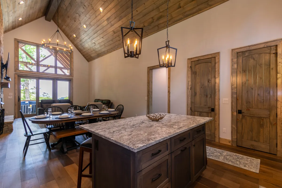 This image captures a welcoming, open-plan interior showcasing the kitchen island, a spacious dining table, large windows allowing natural light, wooden beams, and elegant light fixtures. The layout seamlessly connects the kitchen, dining, and living areas, perfectly illustrating the heart of the home.