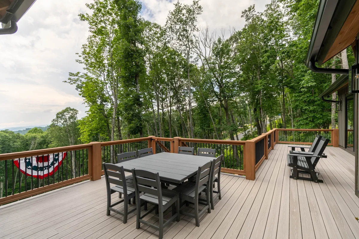 This exterior image features an inviting dining table set on a spacious deck surrounded by lush trees and a sturdy railing. The deck is designed for relaxation and social gatherings, offering an ideal spot for al fresco meals or quiet moments with nature. The natural setting hints at the tranquility and privacy the property provides.